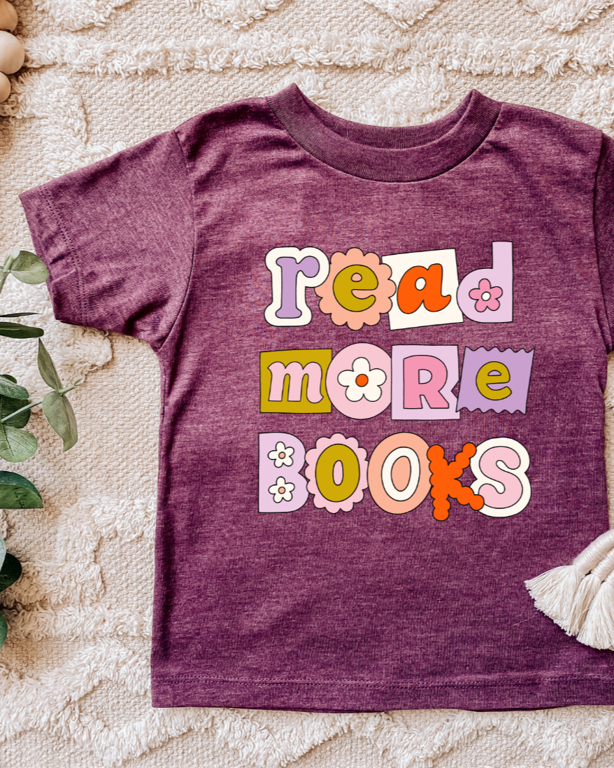 A purple toddler t-shirt with the phrase 'read more books' printed in colorful letters, displayed on a neutral background with some decorative elements.