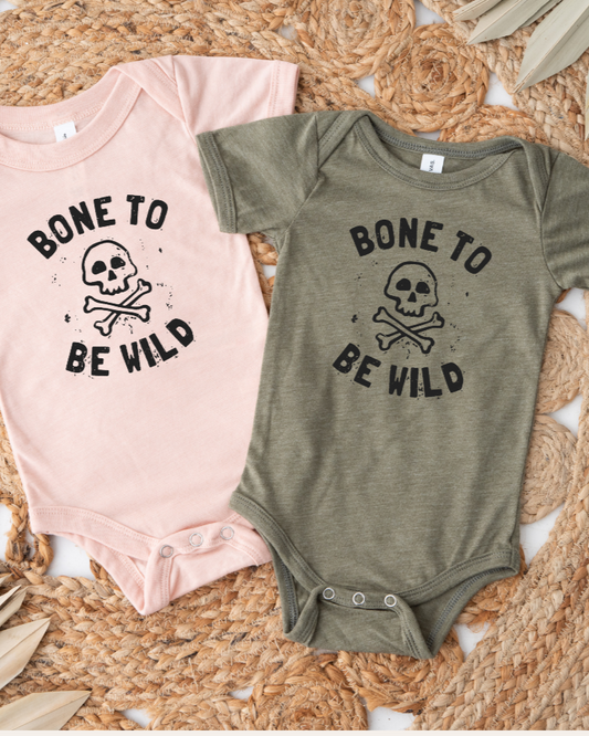 Two baby onesies, one pink and one green, with 'Bone to Be Wild' text and skull design on a woven surface.